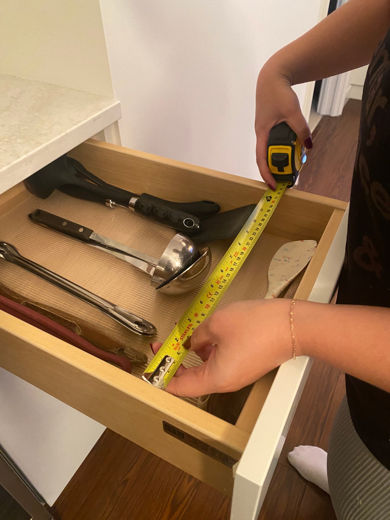 An open drawer with kitchen utensils and a woman's hands using a measuring tape to measure the width of the drawer
