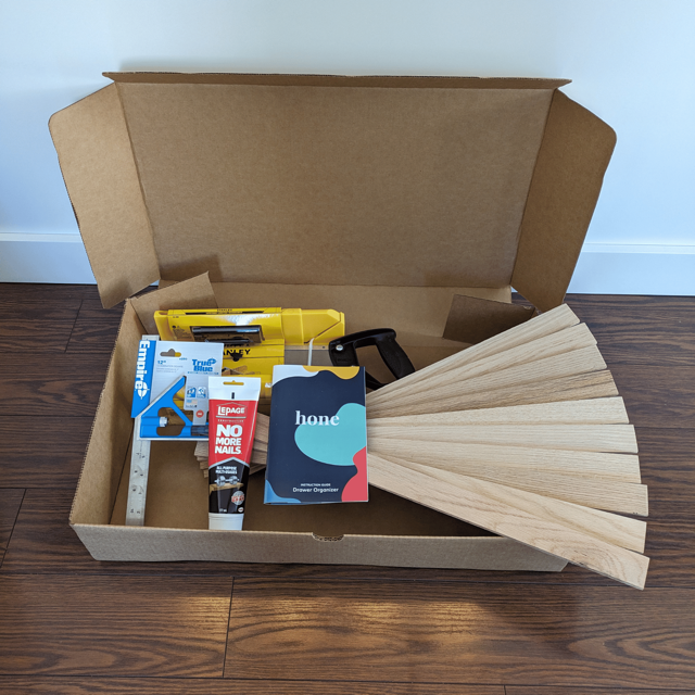 Drawer organizer DIY supplies in an open cardboard box. Box is holding square tool, adhesive glue, Hone instruction guide, oak wood boards, and yellow hand saw.